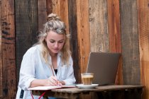 Jovem loira sentada sozinha em uma mesa de café com um computador portátil, escrevendo em livro de anotações, trabalhando remotamente. — Fotografia de Stock