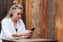 Junge blonde Frau sitzt allein in einem Café, benutzt Handy, arbeitet aus der Ferne. — Stockfoto