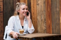 Jeune femme blonde assise seule dans un café, utilisant un téléphone portable, travaillant à distance. — Photo de stock