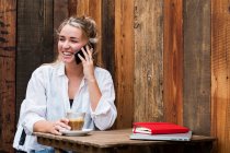 Mujer rubia joven sentada sola en un café, usando teléfono móvil, trabajando remotamente. - foto de stock