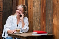 Mujer rubia joven sentada sola en un café, usando teléfono móvil, trabajando remotamente. - foto de stock