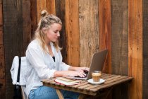 Jeune femme blonde portant un masque facial assise seule à une table de café avec un ordinateur portable, travaillant à distance. — Photo de stock