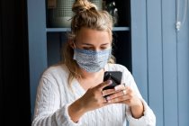Young blond woman wearing face mask sitting alone in a cafe, using mobile phone, working remotely. — Stock Photo