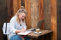 Young blond woman sitting alone at a cafe table with a laptop computer, writing in note book, working remotely. — Stock Photo