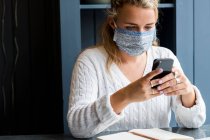 Jeune femme blonde portant un masque facial assise seule dans un café, utilisant un téléphone portable, travaillant à distance. — Photo de stock
