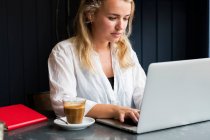 Jeune femme blonde assise seule à une table de café avec un ordinateur portable, travaillant à distance. — Photo de stock