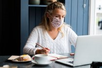 Junge blonde Frau mit Gesichtsmaske sitzt allein an einem Cafétisch mit Laptop, schreibt Notizbuch, arbeitet aus der Ferne. — Stockfoto