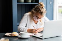 Jeune femme blonde portant un masque facial assise seule à une table de café avec un ordinateur portable, écrivant dans un carnet de notes, travaillant à distance. — Photo de stock