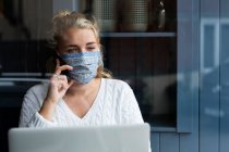 Young blond woman wearing face mask sitting alone at a cafe table with a laptop, using mobile phone, working remotely. — Stock Photo
