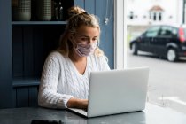 Mujer rubia joven con máscara facial sentada sola en una mesa de café con un ordenador portátil, trabajando remotamente. - foto de stock