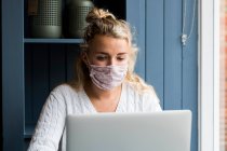 Jeune femme blonde portant un masque facial assise seule à une table de café avec un ordinateur portable, travaillant à distance. — Photo de stock