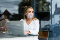 Giovane donna bionda che indossa maschera facciale seduta da sola a un tavolo da caffè con un computer portatile, lavorando a distanza. — Foto stock