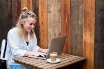 Молода блондинка сидить наодинці за столом кафе з комп'ютером для ноутбуків, працює дистанційно . — стокове фото