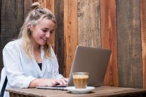 Mujer rubia joven sentada sola en una mesa de café con una computadora portátil, trabajando remotamente. - foto de stock