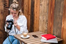 Jeune femme blonde seule à une table extérieure, regardant l'affichage de l'appareil photo numérique. — Photo de stock