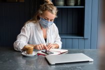 Жінка носить маску для обличчя наодинці за столом кафе з ноутбуком і блокнотом, що працює віддалено . — стокове фото
