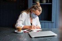 Mujer usando mascarilla solo en una mesa de café con un ordenador portátil y portátil trabajando de forma remota. - foto de stock