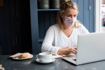 Donna che indossa maschera facciale seduta da sola a un tavolo da caffè con un computer portatile, scrivere nel taccuino, lavorare a distanza. — Foto stock