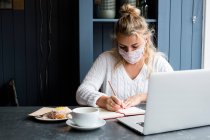 Mujer con máscara facial sentada sola en una mesa de café con un portátil, escribiendo en un cuaderno, trabajando remotamente. - foto de stock