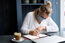 Femme portant un masque facial assis seul à une table de café avec un ordinateur portable, travaillant à distance. — Photo de stock