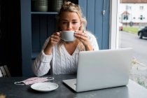 Young blond woman sitting alone at a cafe table with a laptop, and cup of coffee — Stock Photo