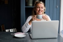 Jovem loira sentada sozinha em uma mesa de café com um laptop e xícara de café — Fotografia de Stock