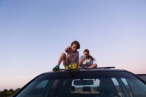 Adolescente menina e seu irmão mais novo sentado no topo do carro SUV ao pôr do sol. — Fotografia de Stock