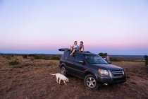 Adolescente menina e seu irmão mais novo sentado no topo do carro SUV ao pôr do sol. — Fotografia de Stock