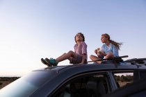 Teenagermädchen und ihr jüngerer Bruder sitzen bei Sonnenuntergang auf SUV-Auto. — Stockfoto