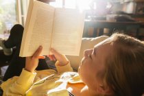 Adolescente leyendo libro en casa en la luz de la mañana temprano - foto de stock