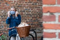 Young blond woman standing next to bicycle with basket, putting on face mask. — Stock Photo
