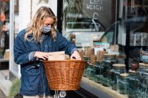 Giovane donna bionda che indossa maschera viso con la bicicletta, in piedi fuori rifiuti libero negozio di alimenti integrali. — Foto stock