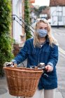Young blond woman wearing face mask on bicycle with basket, looking at camera. — Stock Photo