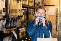 Молодая блондинка в маске для лица, делает покупки в бесплатном магазине цельной пищи. — стоковое фото