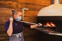 Блондинка в синем полосатом фартуке и маске, стоящая у печи для пиццы в ресторане. — стоковое фото