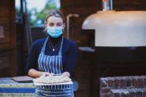 Woman waitress in apron and face mask holding fresh pizza on a board — Stock Photo