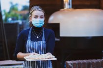 Жінка-офіціантка в фартусі та масці для обличчя, що тримає свіжу піцу на дошці — стокове фото