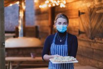 Жінка-офіціантка в фартусі та масці для обличчя, що тримає свіжу піцу на дошці — стокове фото