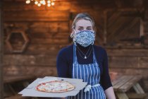 Жінка-офіціантка в фартусі та масці для обличчя, що тримає тарілку піци . — стокове фото