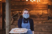 Mujer camarera en delantal y máscara facial sosteniendo plato de pizza. - foto de stock