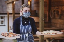 Woman waitress in apron and face mask holding plates of pizza. — Stock Photo