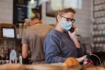 Garçonete loira usando máscara facial trabalhando em um café, no telefone. — Fotografia de Stock