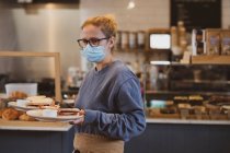 Cameriera bionda che indossa maschera viso lavorando in un caffè, portando piatti di cibo. — Foto stock