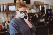 Blonde Kellnerin mit Mundschutz bei der Arbeit in einem Café, am Telefon. — Stockfoto