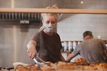 Блондинка офіціантка носить маску для обличчя, що працює в кафе, розмиваючи випічку . — стокове фото