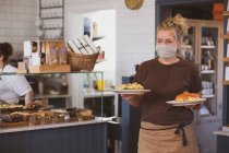 Блондинка офіціантка носить маску для обличчя, що працює в кафе, несучи тарілки їжі . — стокове фото