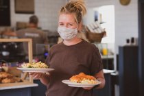 Blonde Kellnerin mit Mundschutz arbeitet in einem Café und trägt Teller mit Lebensmitteln. — Stockfoto