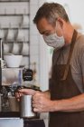 Homem barista vestindo avental marrom e máscara facial trabalhando em um café, espuma de leite. — Fotografia de Stock