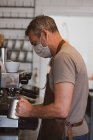 Barista homme portant tablier marron et masque facial travaillant dans un café, moussant lait. — Photo de stock