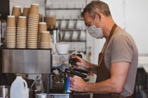 Чоловіча бариста в коричневому фартуху та масці для обличчя, що працює в кафе, робить еспресо . — стокове фото